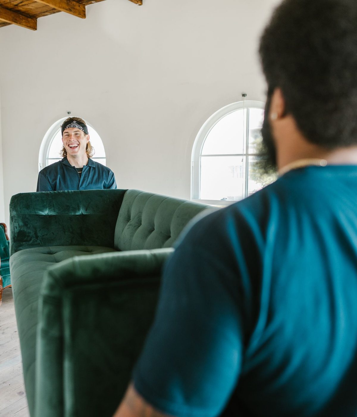Two delivery crew members carrying a green velvet sofa into a bright, freshly moved-into room.