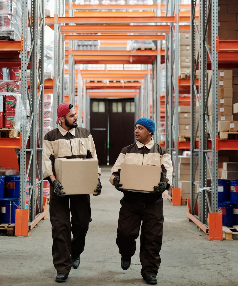 Two warehouse workers in uniform carrying boxes down an aisle of tall storage racking.